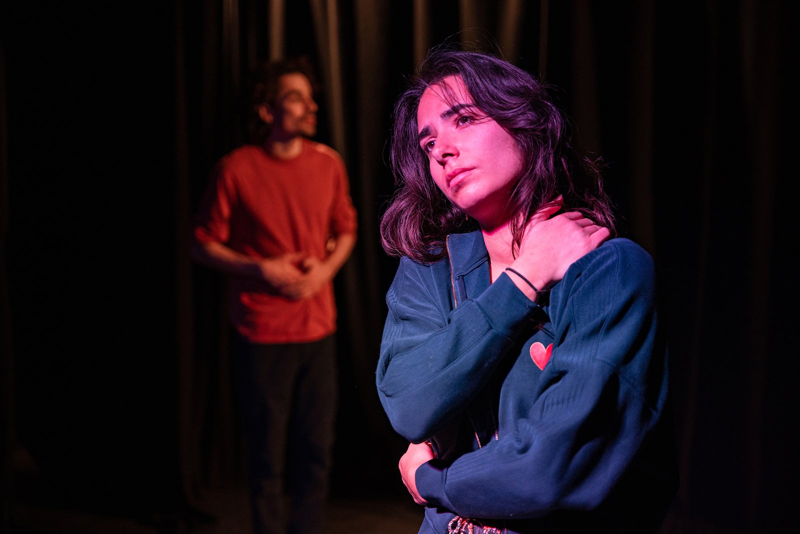 Comédienne au premier plan en pleine réflexion pendant qu’un autre personnage apparaît en arrière-plan sur scène.